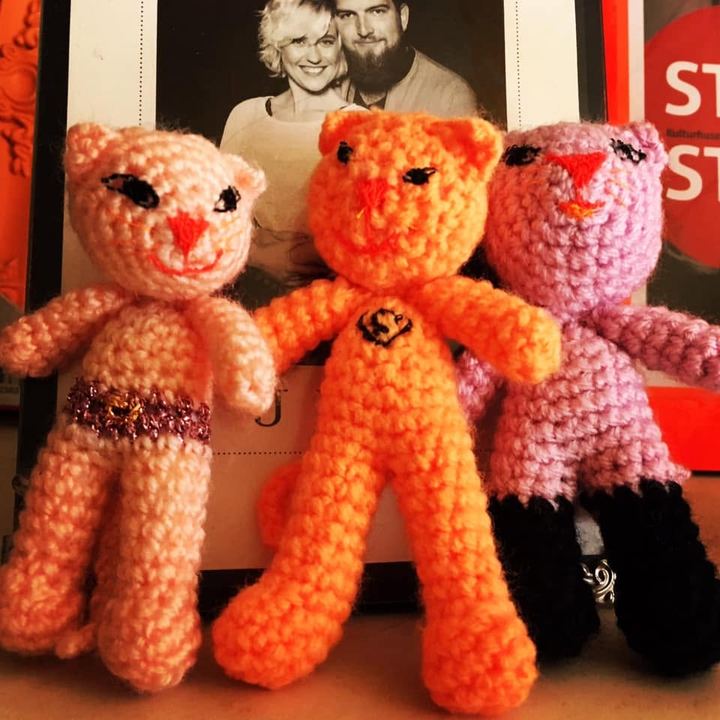 Tre hæklede katte i lyserød, orange og rosa garn står på række med brede smil.